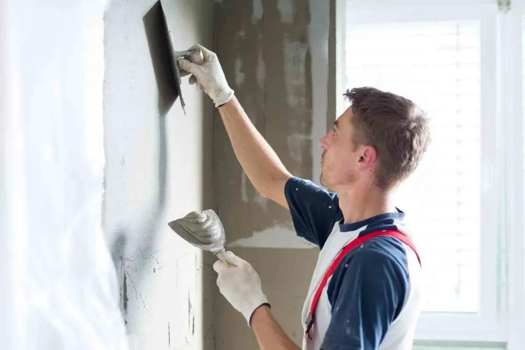 A worker in gloves applying plaster to an interior wall with a trowel and a hawk.
