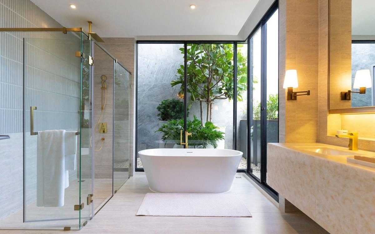 A modern bathroom with a freestanding tub, glass shower enclosure, brass fixtures and a planted courtyard beyond floor-to-ceiling windows.